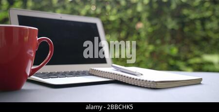 Computer laptop with notepad and coffee in red cup with green nature background. Work remotely or from home. Copy space. Stock Photo