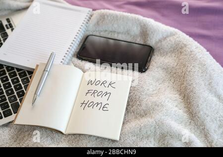 Work from home, text written on notepad. Computer laptop, cellphone and notepad on top of bed in the bedroom. Stock Photo