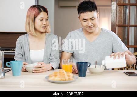 happy asian couple in pajamas sitting at table in kitchen at home in morning and having cereal breakfast together Stock Photo