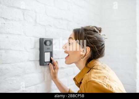 Woman rings the house intercom with a camera installed on the white brick wall Stock Photo