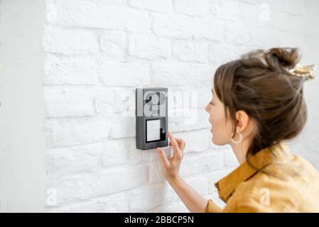 Woman rings the house intercom with a camera installed on the white brick wall Stock Photo