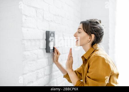 Woman rings the house intercom with a camera installed on the white brick wall Stock Photo