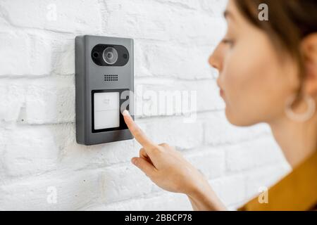 Woman rings the house intercom with a camera installed on the white brick wall Stock Photo