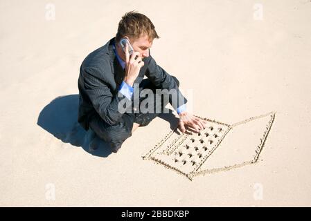 Telecommuting businessman multitasking between talking on his mobile phone and typing on a laptop computer hand drawn in smooth sand on a sunny beach Stock Photo