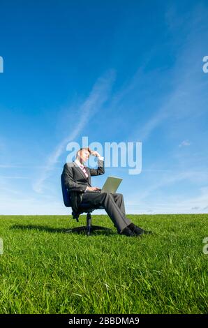 Thinking businessman sitting outdoors on an office chair working on a laptop scratching his head looking up in a sunny green meadow Stock Photo