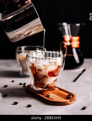 https://l450v.alamy.com/450v/2bbeah9/the-process-of-making-iced-coffee-ice-cube-in-a-glass-pouring-milk-wooden-tray-scattered-grains-concrete-background-2bbeah9.jpg