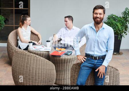 Prtrait of happy confident bearded entreprneur leaning on wicker cafe chair and looking at camra, his colleagues talking in background Stock Photo