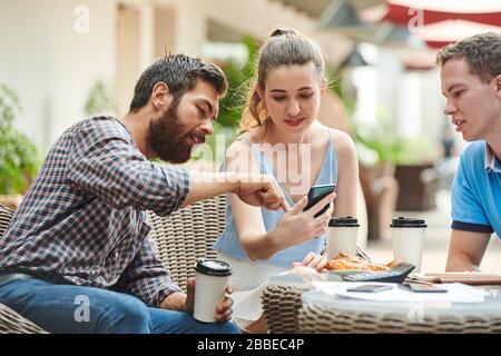 Group of coworkers having lunch in outdoors cafe and discussing new mobile application on smartphone screen Stock Photo