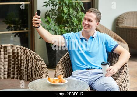 Handsome smiling young man taking selfie in outdoor cafe when having coffee with croissants for breakfast Stock Photo