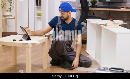 Male worker in overalls assembles new white furniture in new home owners. Man reading instruction on laptop. Stock Photo