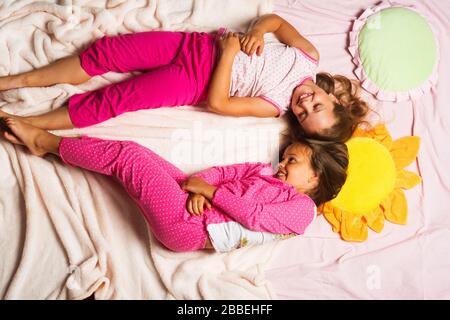 Kids in pink pajamas put heads together. Children with happy faces lie close on light pink blanket background. Schoolgirls having pajama party with funny pillows. Childhood and happiness concept Stock Photo