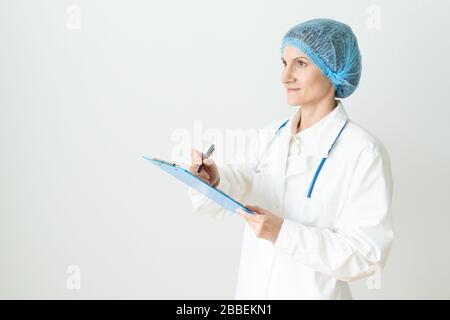 Female adult doctor stands on white background. Beautiful woman, thinking, smiling, writing. Clipboard in his hand, stethoscope around his neck . Stock Photo
