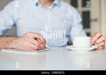 Man writes to-do list in notebook, working at home. Drink coffee, keep a pencil. Wearing blue casual shirt. No gadgets Stock Photo
