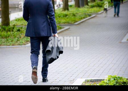 Walking businessman on sidewalk in a park. Businessman in blue business suit holding a jacket. Stock Photo