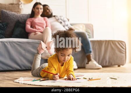 Child draws on the floor, parents sitting on couch Stock Photo