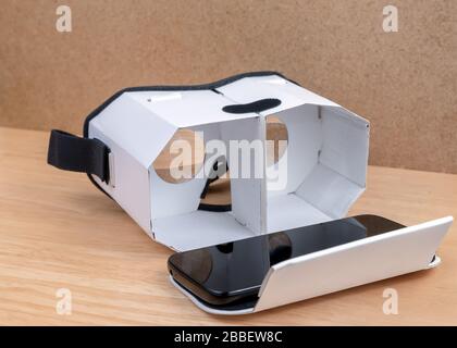 A cardboard virtual reality headset. Front view, it is open a mobile phone on it. Lenses visible inside. It is resting on a wooden table next to a cor Stock Photo