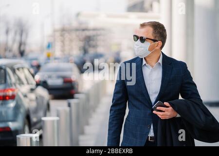 Thoughtful entrepreneur in formal wear, sunglasses, protects himself with sterile medical mask against coronavirus disease, walks outdoor, holds mobil Stock Photo