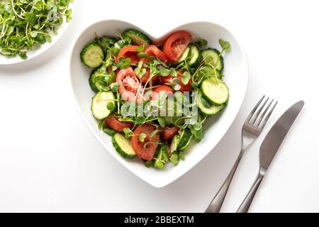 Fresh salad with tomato, cucumber, vegetables, microgreen radishes in plate shape of heart on white. Concept vegan and healthy eating. Top view. Stock Photo