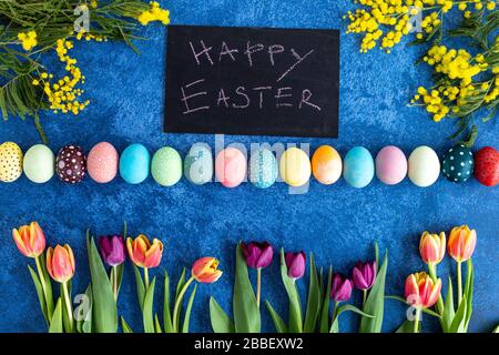 Colorful Easter eggs and tulips on dark blue background. On blackboard is written happy easter. Holiday decorations, Easter concept background. Stock Photo