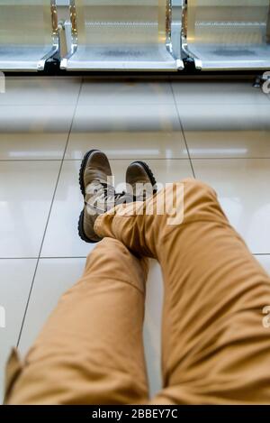Man sitting with his legs in hiking boots on floor in the airport parking lot waiting for his connection. Stock Photo