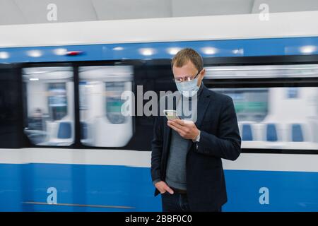 Horizontal shot of male worker poses at subway platform, commutes by public transport, uses modern mobile phone to check route, wears medical protecti Stock Photo