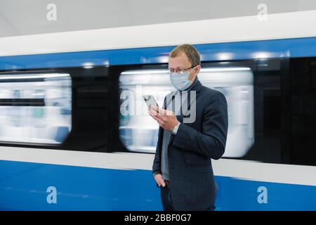 Horizontal shot of male traveler uses public transport for commuting, wears medical mask to protect from coronavirus or covid-19, waits for train, use Stock Photo
