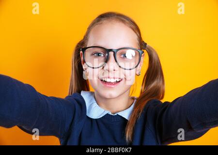 Close up of little kid girl 12-13 years old in glasses doing selfie on mobile phone on yellow background. Stock Photo