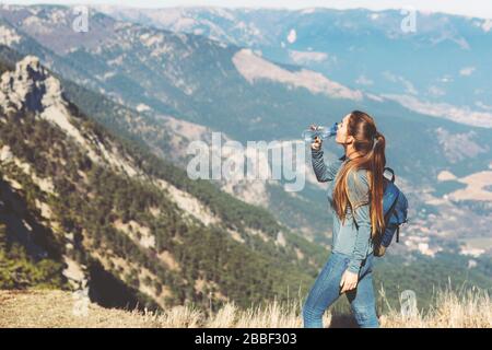 Young beautiful girl travels alone in the mountains in spring or autumn, looks into the distance and enjoys nature, rocks and green forests, view of the landscape. a backpack behind and sportswear, drinking water from an eco bottle Stock Photo