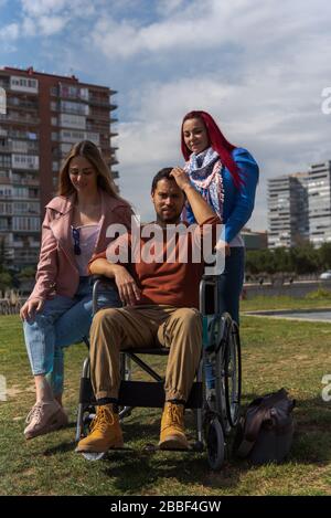 Young Latino paraplegic in a wheelchair in a worried park with two young Caucasian girls with buildings in the background Stock Photo