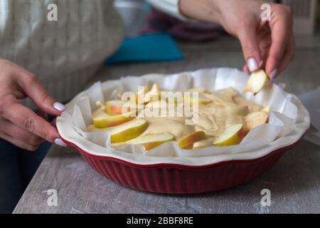 The hostess at home is preparing apple charlotte in the kitchen. puts a slice of apple on a pie in a baking dish. Raw Apple Pie Dough Stock Photo