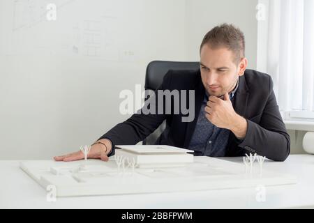 Businessman in office sitting at desk looking over a house model. Thoughtful architect thinking about project. Stock Photo