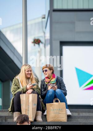 Two friends take a seated rest and share a joke whilst shopping at Westfield Shopping Centre, Stratford. Complete with shopping bags and phones. Stock Photo