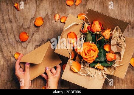 Woman's hands open craft paper envelope next to open box with orange roses in it, close up. Gifts concept. Wooden background with rose petals, top view. Stock Photo
