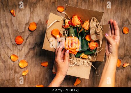 Woman's hands hold open box with orange roses in it, close up. Gifts concept. Wooden background with rose petals and craft paper envelope, top view. Stock Photo