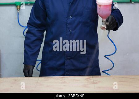 A young man in a dark coat applies a glue on a wooden piece of the sofa, on a working desk. Behind him are a lot of tools and materials. He is using a Stock Photo