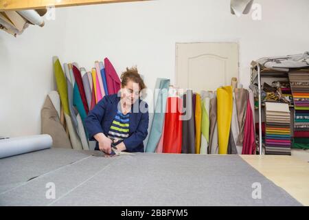 An older woman in a dark coat cuts the marked grey material for the sofa. Behind the are different types of materials. She is cutting the material on Stock Photo