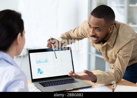 Cheerful young Afro-American manager pointing at online graphs on laptop while offering business solution Stock Photo
