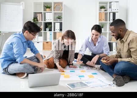 Group of positive young multi-ethnic specialists sitting on floor and working on startup project together Stock Photo