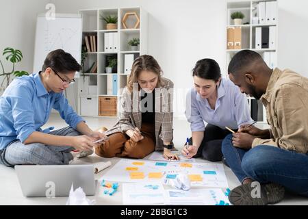 Group of interracial startuppers sitting on floor with task board and analyzing marketing strategies in office Stock Photo