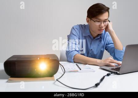 Pensive young Asian analyst in eyeglasses sitting at desk with projector and using laptop while working on presentation Stock Photo