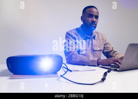 Handsome Afro-American manager sitting at desk and using laptop while checking presentation on projection screen in meeting room Stock Photo