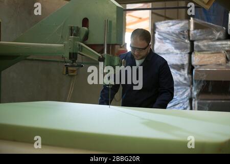 Process of furniture production. A young man in a dark coat cut the foam for the sofa on a cutting machine. Behind him there are a lot of finished sof Stock Photo