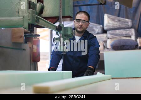 Process of furniture production. A young man in a dark coat cut the foam for the sofa on a cutting machine. Behind him there are a lot of finished sof Stock Photo