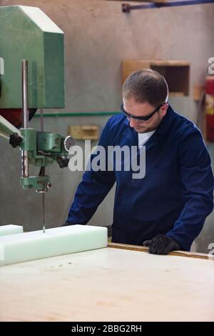 Process of furniture production. A young man in a dark coat cut the foam for the sofa on a cutting machine. He is wearing a safety glasses and gloves. Stock Photo