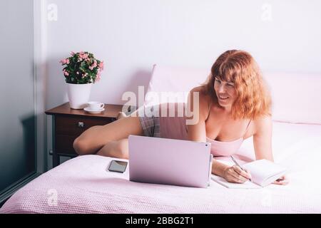 Happy middle age woman working at home on a laptop sitting on the bed in the house. Work from home concept. Self isolation. Stock Photo