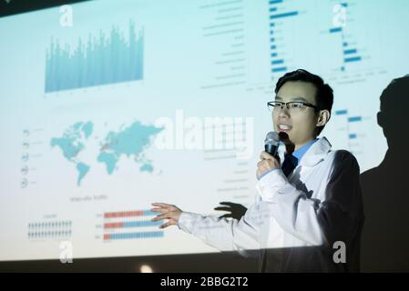 Enterprising young Asian man in glasses and lab coat pointing at projection screen while addressing scientific conference Stock Photo