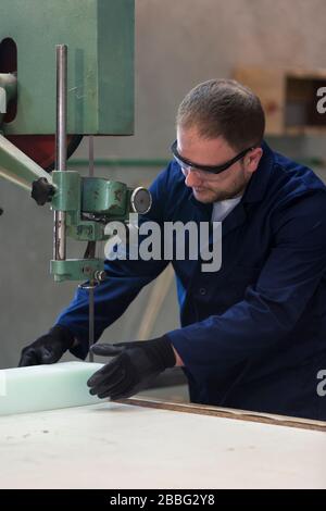 Process of furniture production. A young man in a dark coat cut the foam for the sofa on a cutting machine. He is wearing a safety glasses and gloves. Stock Photo