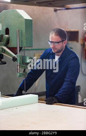 Process of furniture production. A young man in a dark coat cut the foam for the sofa on a cutting machine. He is wearing a safety glasses and gloves. Stock Photo