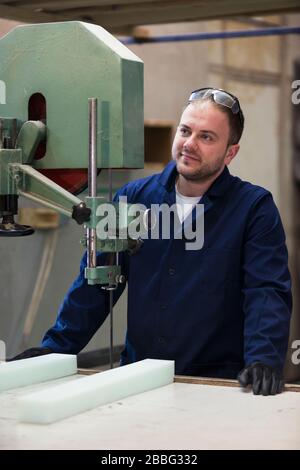 Process of furniture production. A young man in a dark coat cut the foam for the sofa on a cutting machine. He is wearing a safety glasses and gloves. Stock Photo