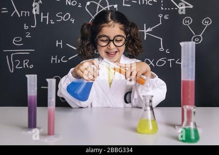 little scientist girl with glasses in lab coat mixing chemical liquids in flasks, on blackboard background with science formulas, concept of back to s Stock Photo